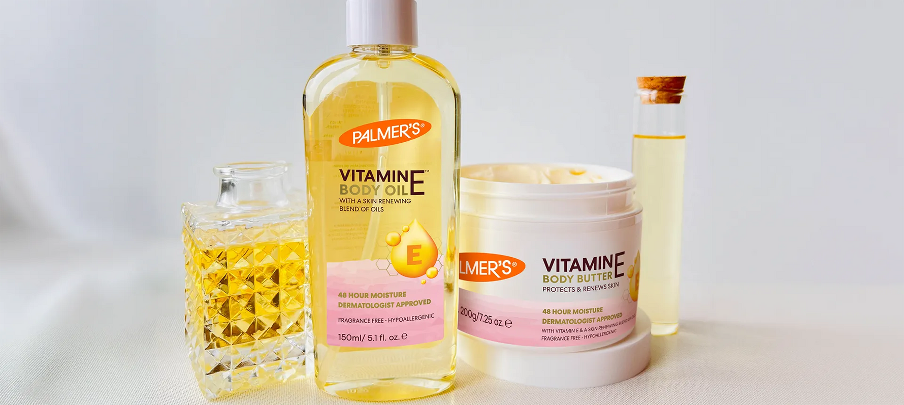 Palmer's Vitamin E Body Oil and Cream on table with beaker and perfume bottle